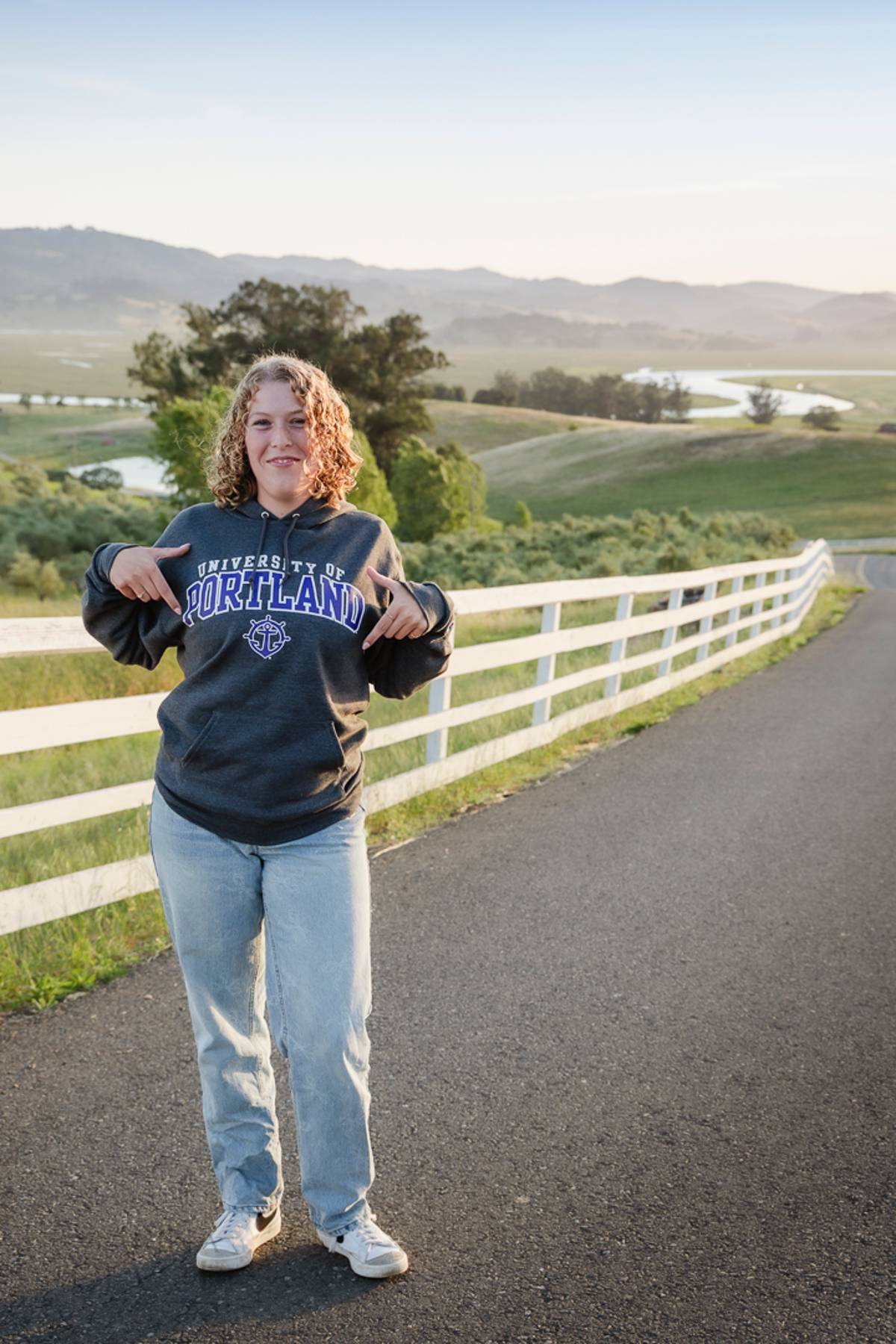 Senior standing on a country road pointing to her college hoodie